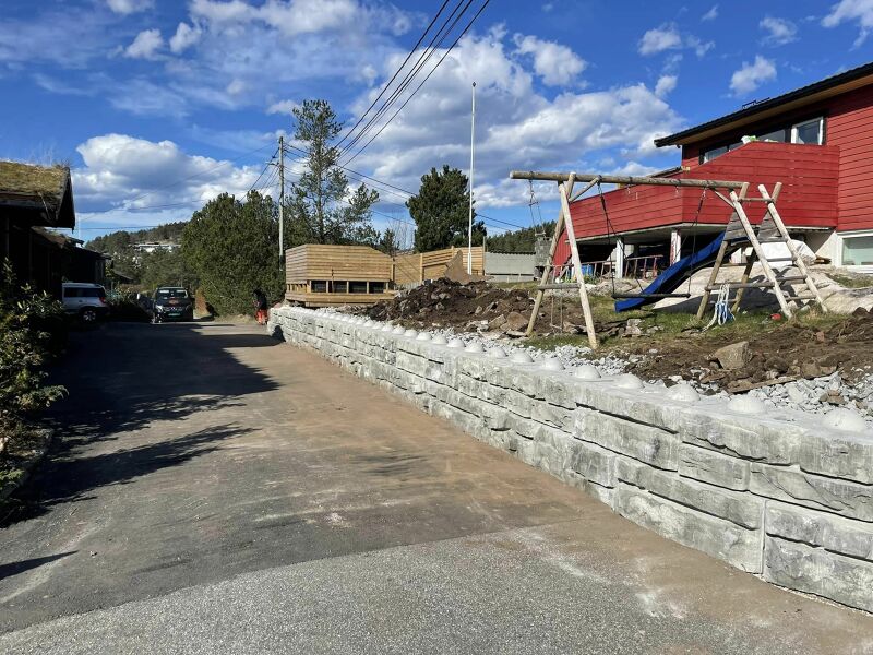Grå støttemur foran rødt hus og lekeplass.